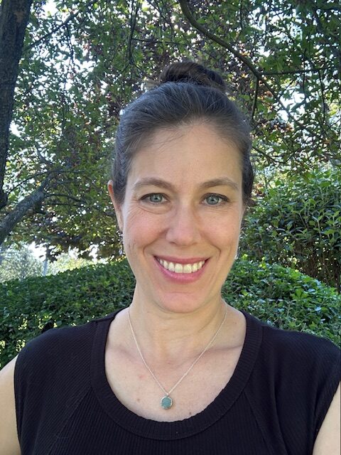 Photo of HTA Practitioner Dana Brady, who has dark hair pulled back into a bun, is wearing a dark shirt, and standing in front of green trees and bushes.