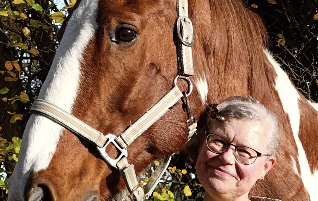 Woman with her brown and white horse whose head is resting on her shoulder from behind as she holds its bridle.